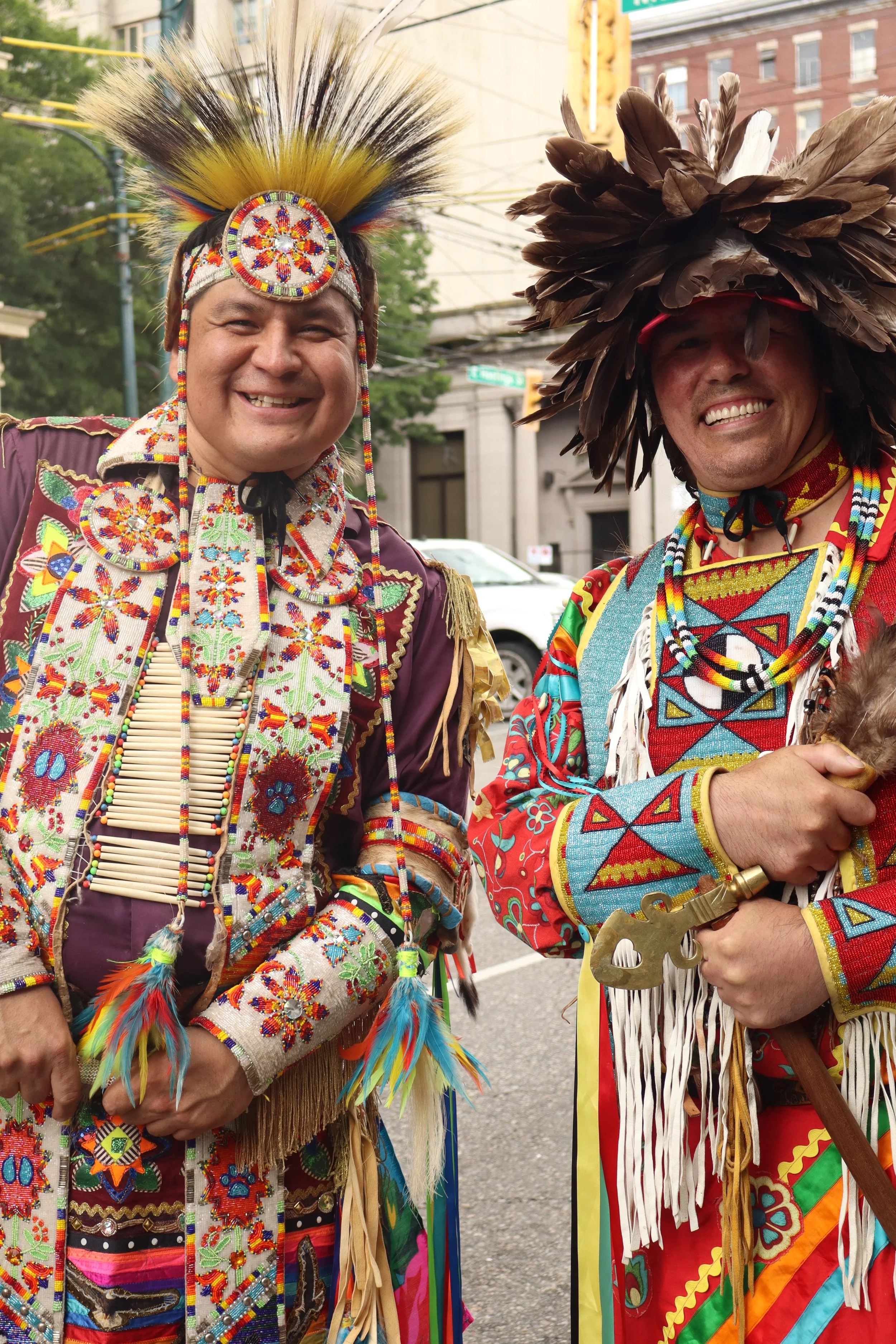 Two people dressed in traditional Native American attire, wearing feathered headdresses and colorful beadwork, smiling at an outdoor event.
