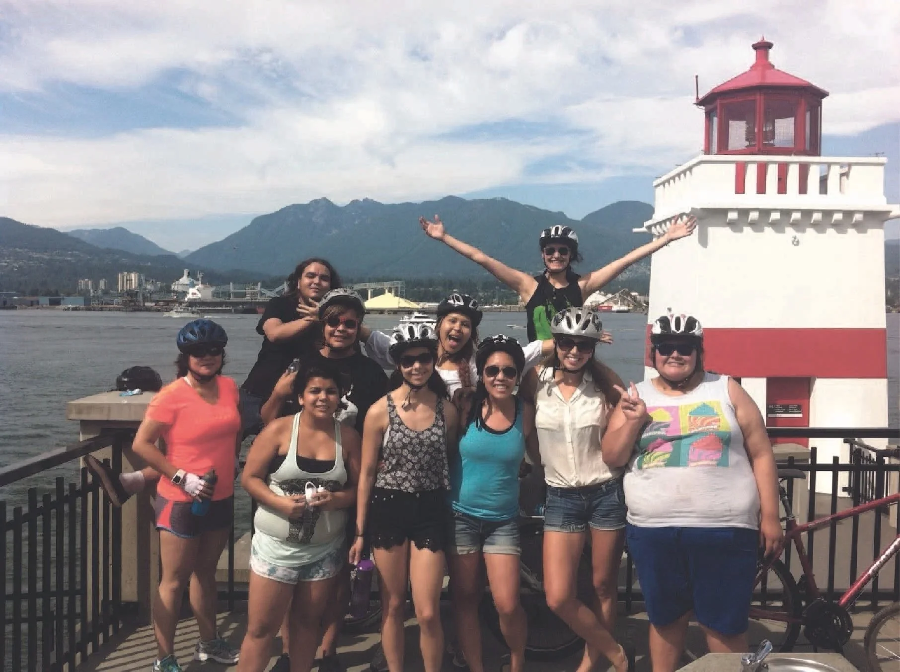 Group of women in cycling gear and helmets posing for photo near a lighthouse by the water with mountains in the background.