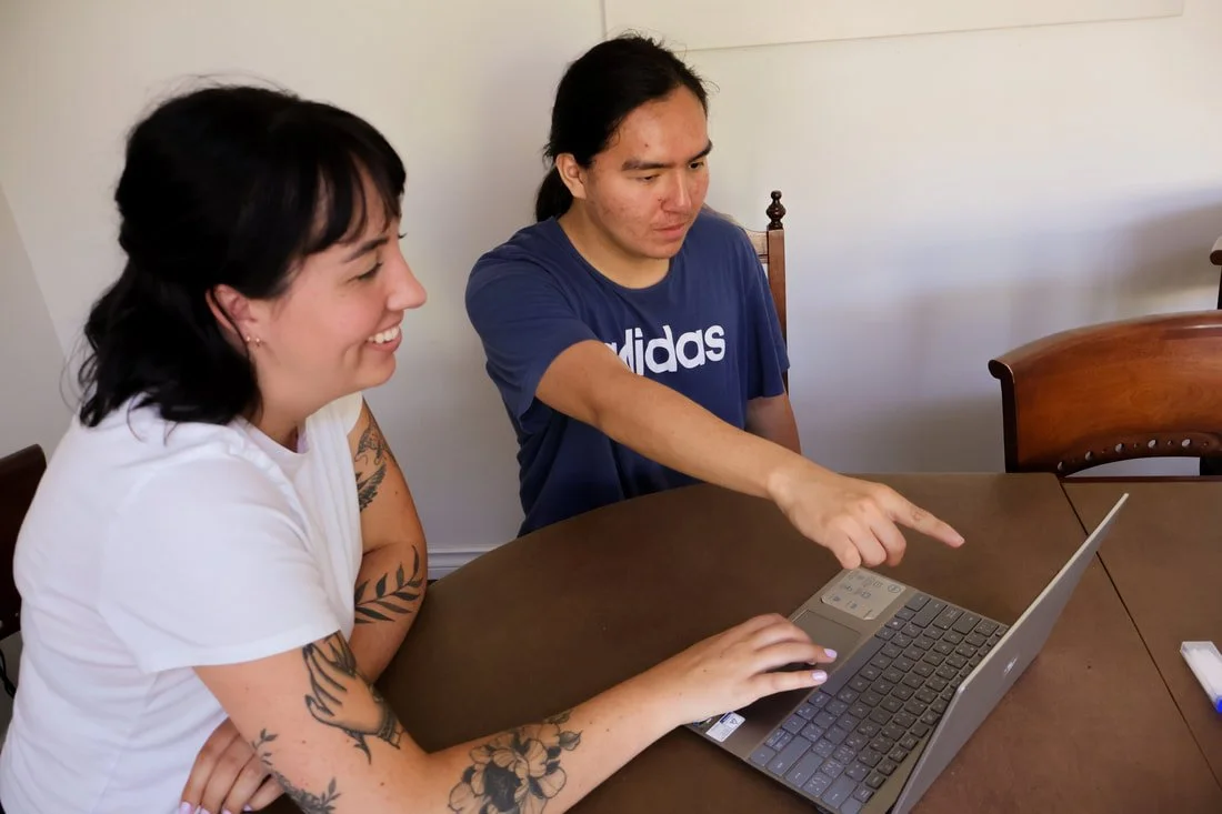 Two women sitting at a wooden dining table, looking at a laptop. One woman is smiling and typing, while the other is pointing at the screen. The woman on the left has tattoos on her arms, and the woman on the right wears a blue Adidas T-shirt.