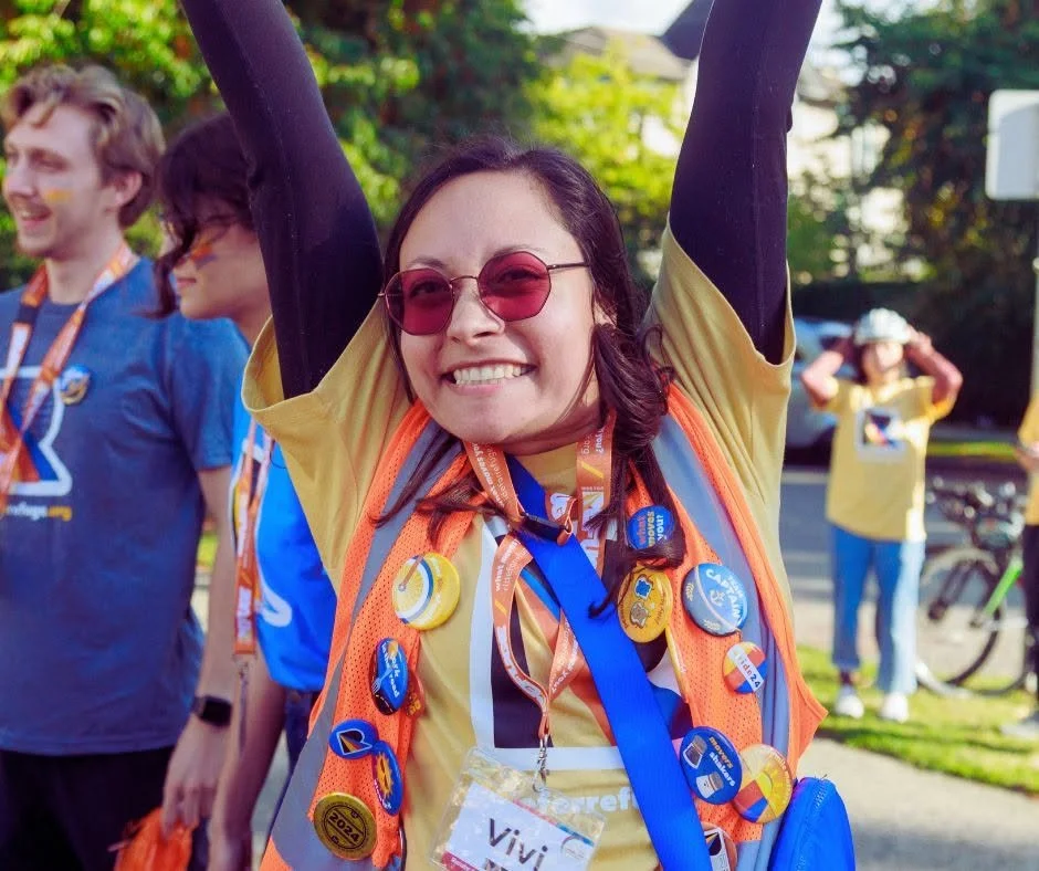 Woman smiling with arms raised, wearing orange vest decorated with medals and badges, sunglasses, and a yellow T-shirt at a cheerful outdoor event, with other participants in the background.