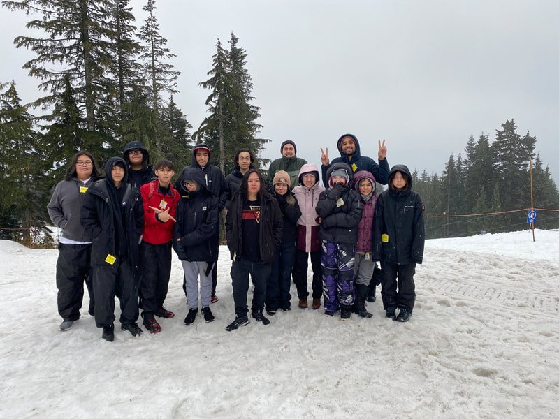 Group of people standing on snow-covered ground in a forested area, some making peace signs, dressed in winter clothing.
