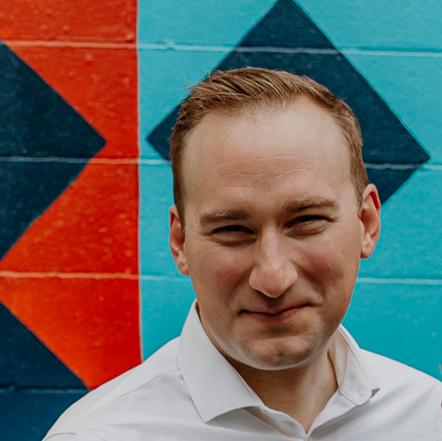 Portrait of a man in a white shirt standing in front of a colorful geometric mural on a brick wall.