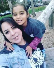 A woman and a young girl are outdoors, smiling and hugging. The woman has dark hair and is wearing a light blue jacket, while the girl has dark hair tied back and is wearing a navy jacket and pink scarf. They are near playground equipment.