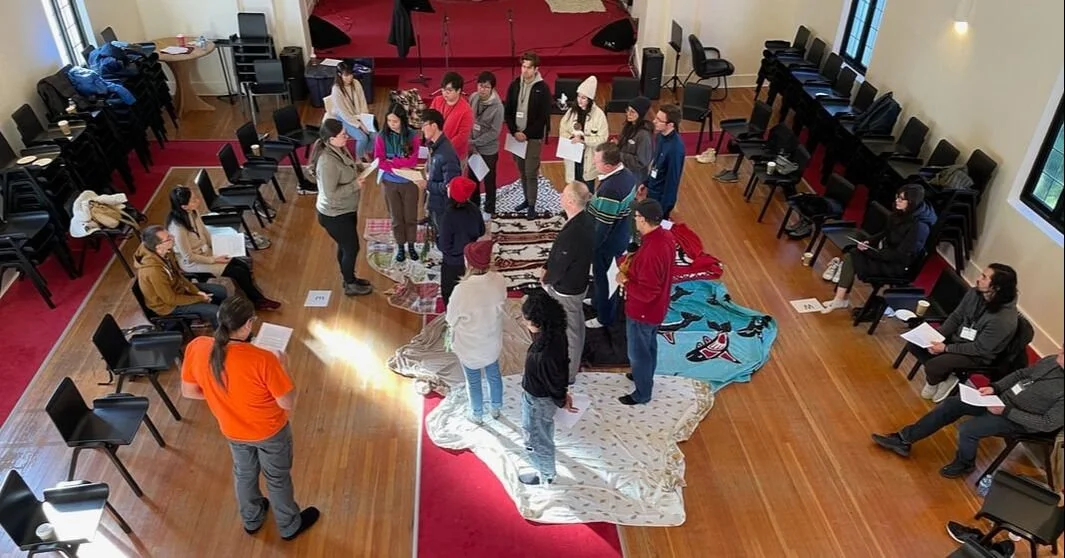 A group of people standing and sitting in a circle in a room with wooden floors, some sitting on chairs around the circle, others standing on fabric and rugs on the floor, with sound equipment, chairs, and windows visible.