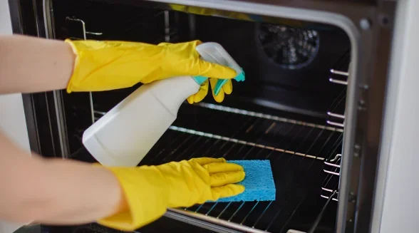 Person cleaning the inside of an oven with a spray bottle and blue cleaning cloth, wearing yellow rubber gloves.
