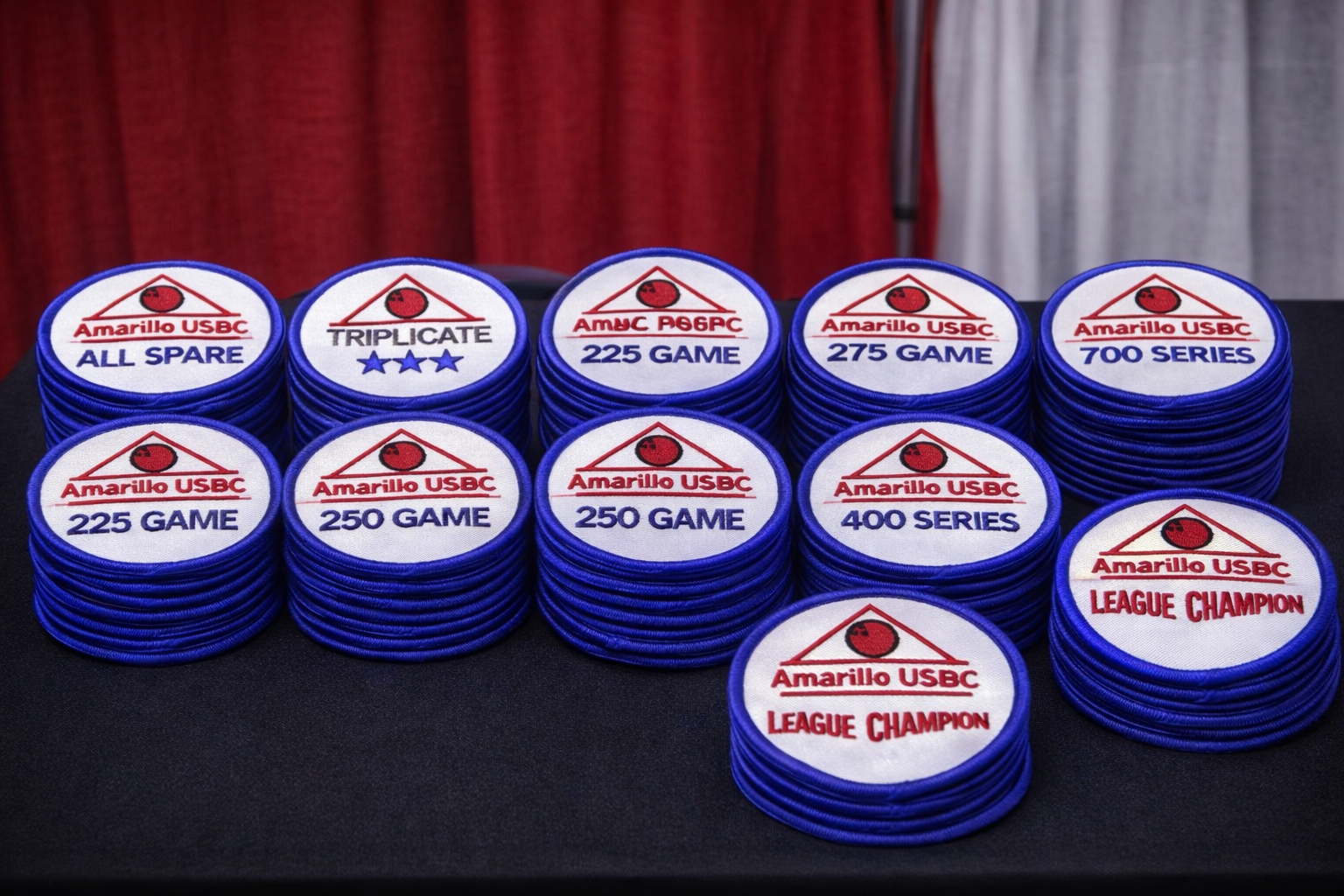 Stacks of embroidered bowling patches with blue borders, featuring various titles related to bowling, such as "ALL SPARE," "TRIPLICATE," and championship titles.