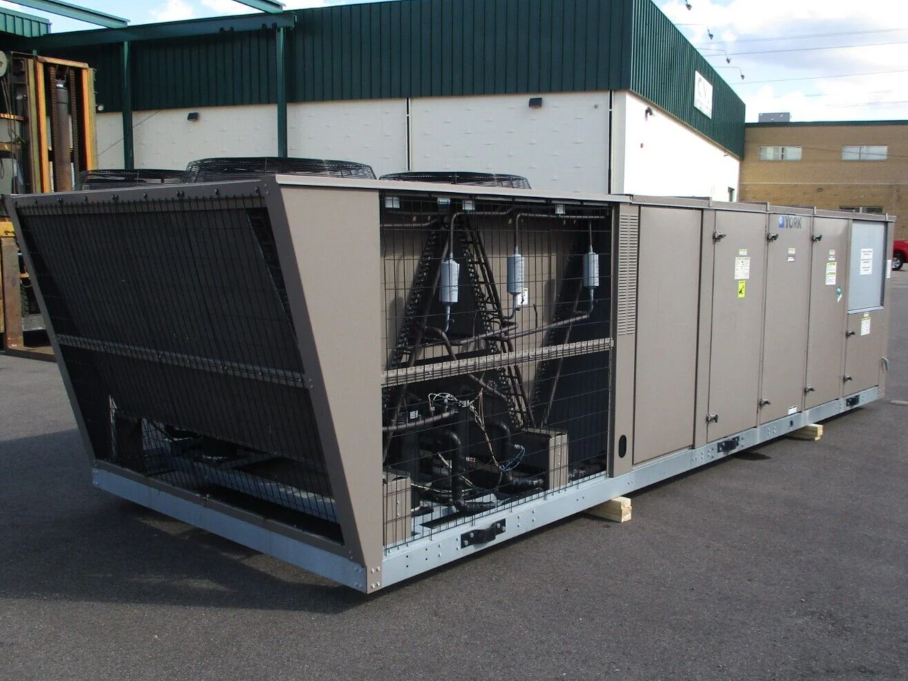 Large industrial HVAC unit with exposed components, situated outdoors on a paved surface near a building with green and white exterior walls.
