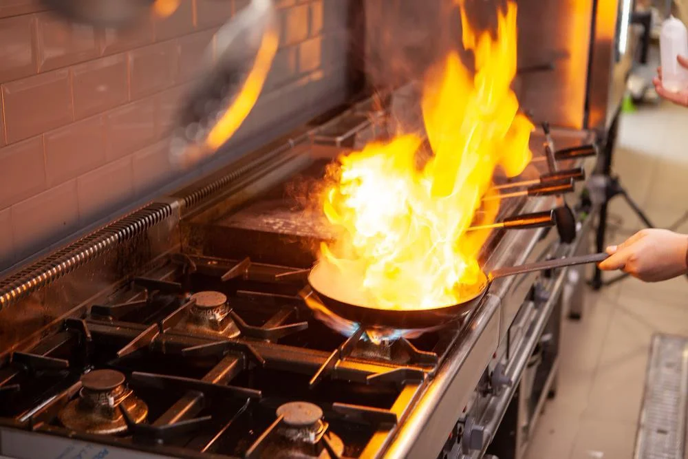 A chef using a pan on a stovetop with flames shooting up during cooking, with a brick wall behind.
