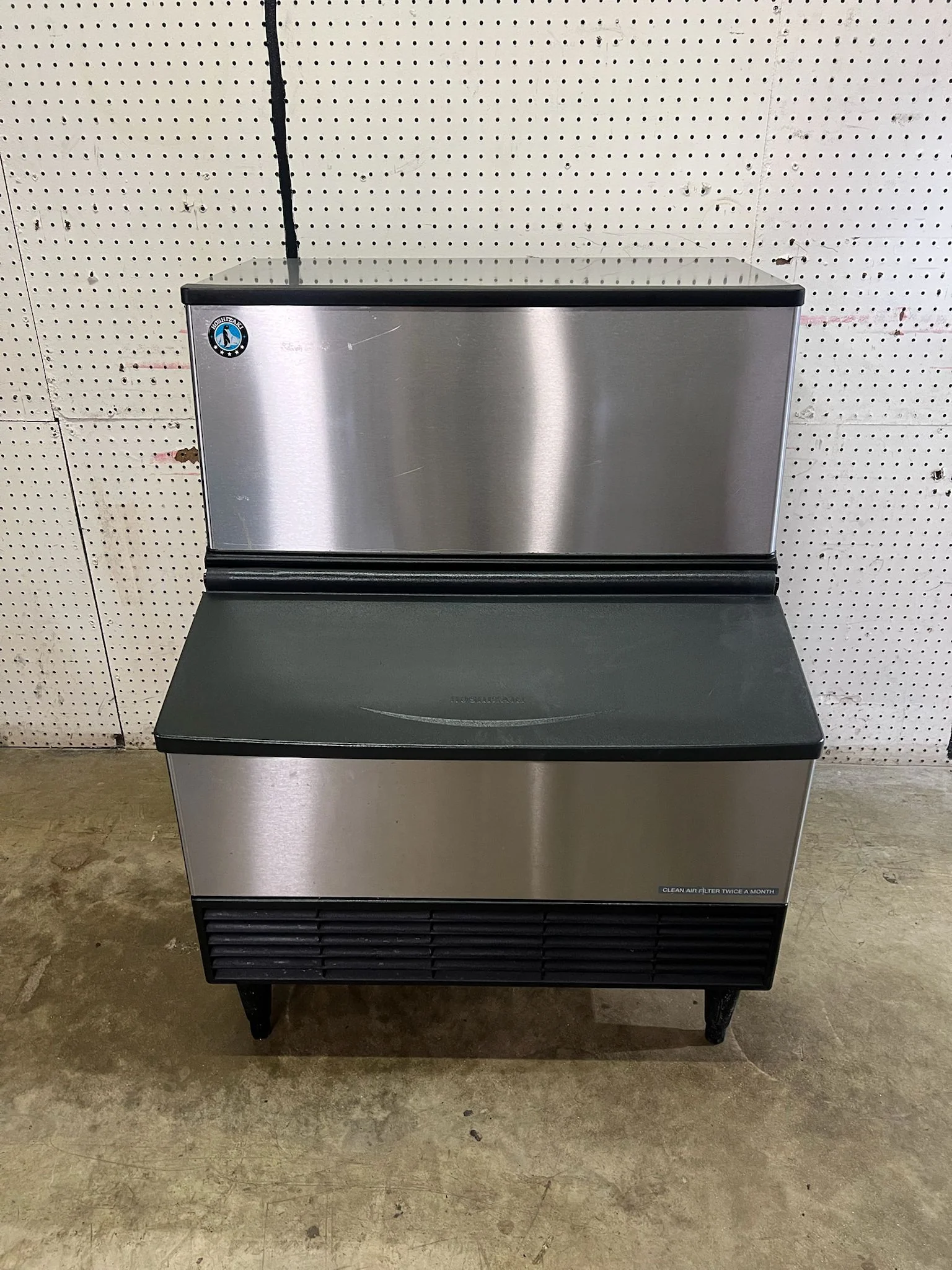 A stainless steel refrigerated display case with a black top and bottom trim, and a small sticker on the upper left corner. The display case is positioned against a pegboard wall with multiple holes, and is sitting on a concrete floor.