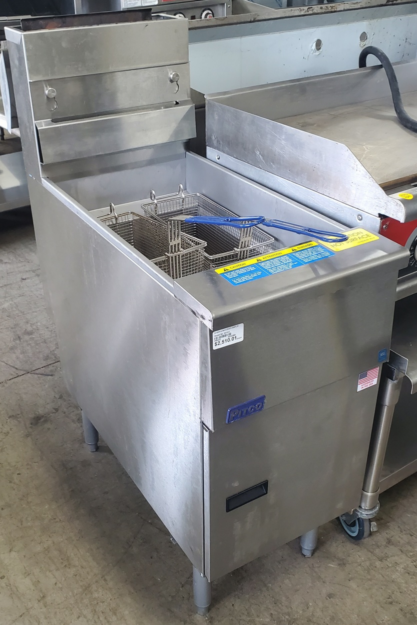 Commercial stainless steel deep fryer with frying baskets and a lid, on a concrete floor in an industrial kitchen setting.