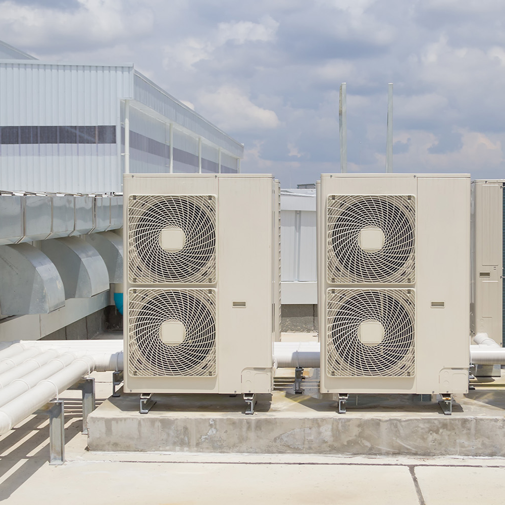 Two HVAC outdoor units on a building rooftop with pipes and a metal railing, against a cloudy sky.