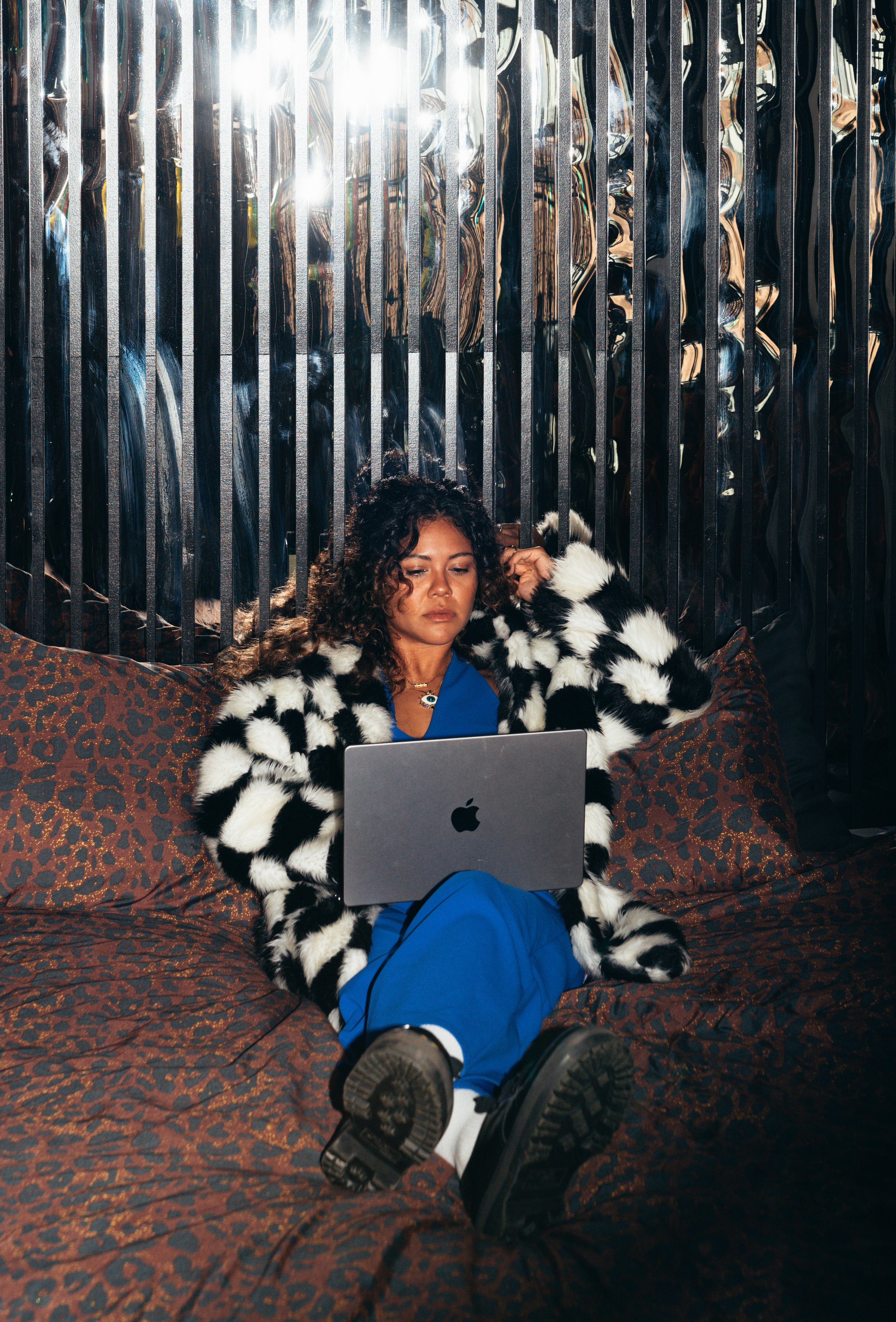 A woman sitting on a leopard print bedspread using a silver MacBook laptop, wearing a black and white fur coat, blue pants, and black boots.