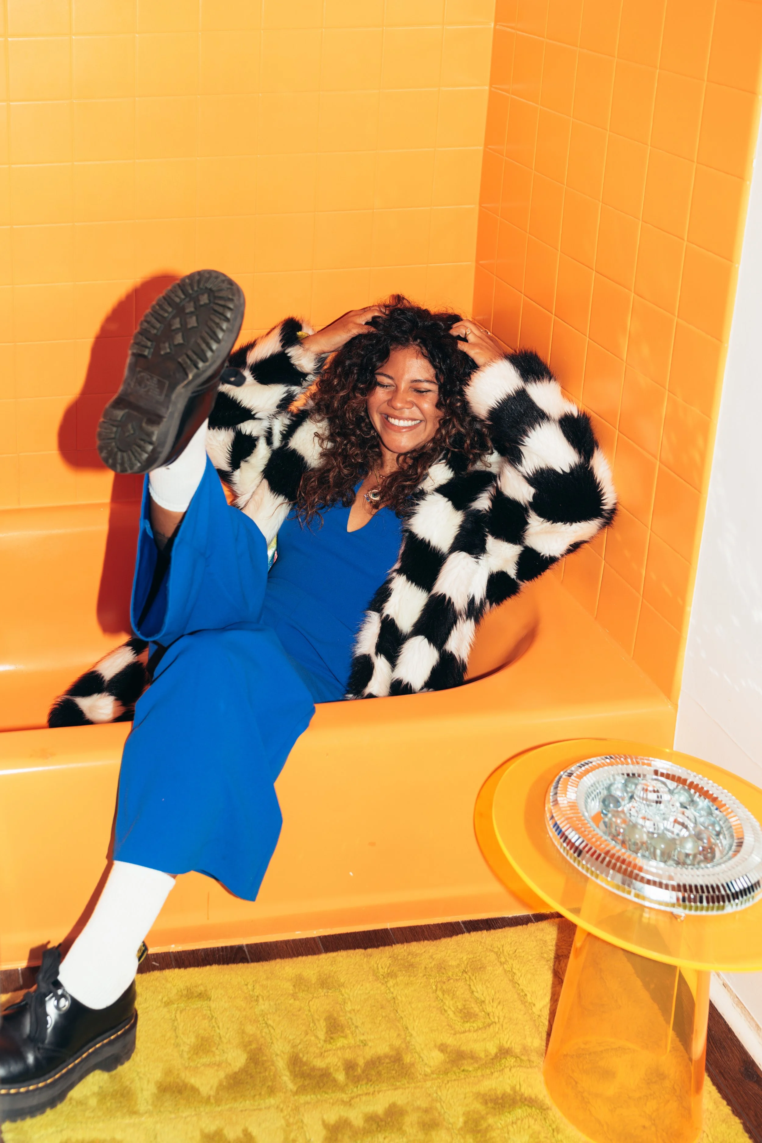 A woman with curly hair smiling and laughing while sitting in an orange bathtub in a brightly colored room with orange tiled walls, wearing a black and white striped fur coat, blue scrubs, white socks, and black shoes.