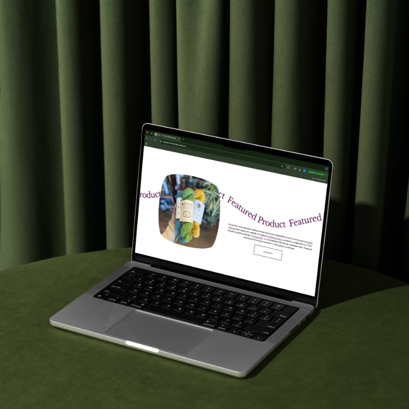 A laptop on a green table showing a website featuring colorful yarn skeins.