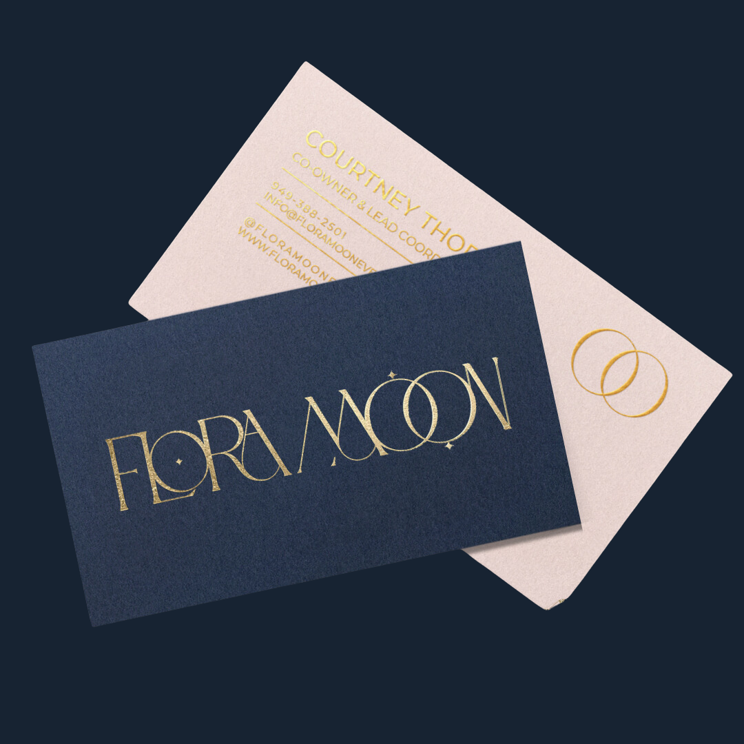 Business cards for Flora Moon, one navy blue with gold lettering, and one light pink with gold rings and contact information.