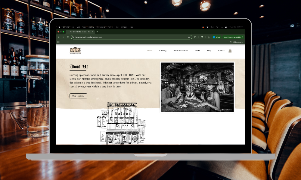 A laptop on a wooden table showing a bar's website with an about us section and a black-and-white photo of people at the bar, in a dimly-lit bar or restaurant setting with bottles on shelves in the background.