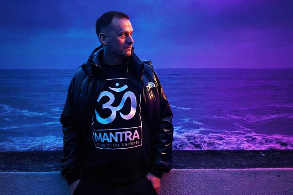 A man stands by the ocean at sunset with a colorful sky in the background. He is wearing a black jacket and a black T-shirt with a white Om symbol and the word 'MANTRA' printed on it.
