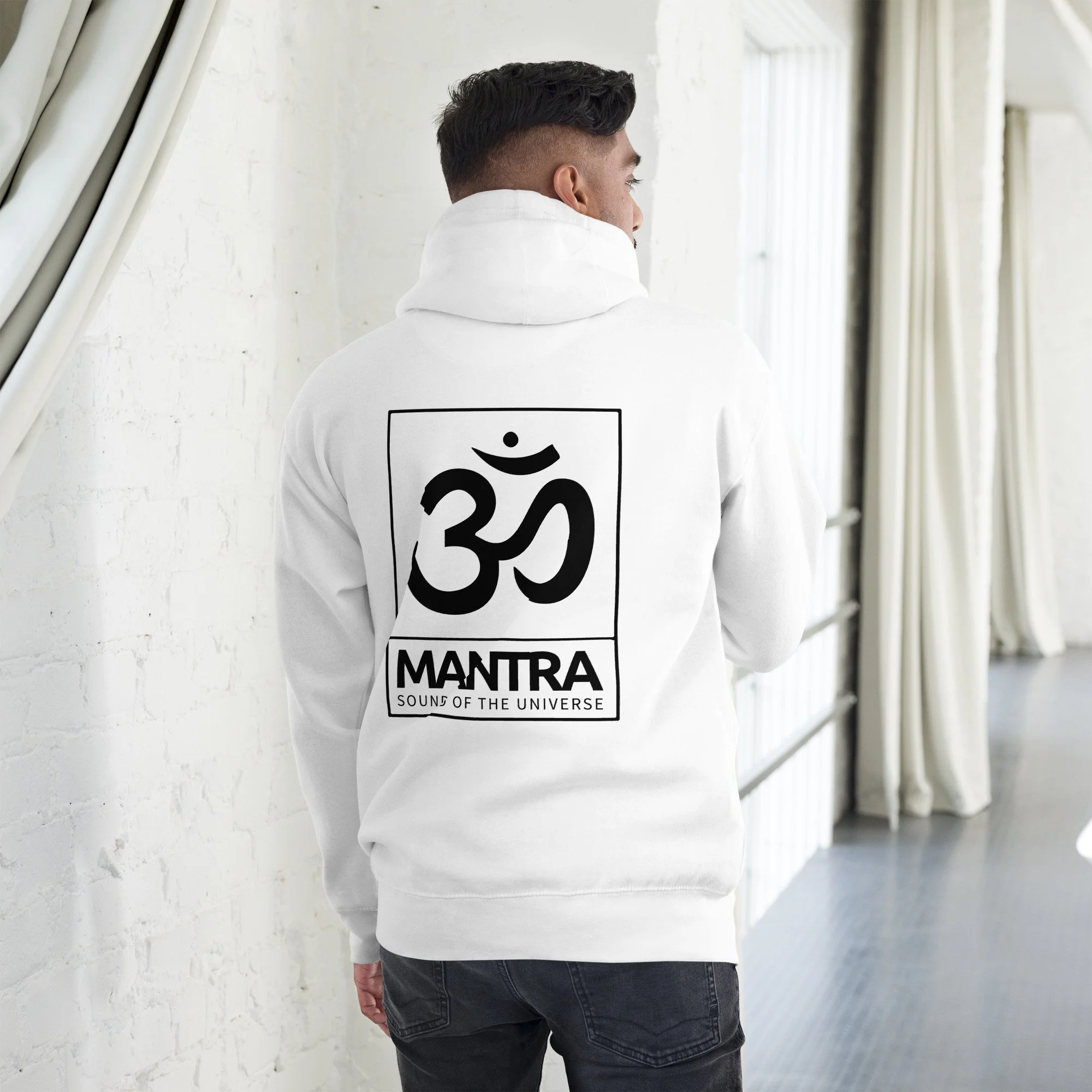 A young man with short dark hair, wearing a white hoodie with a graphic and the words "MANTRA SOUND OF THE UNIVERSE" on the back, is standing with his back to the camera in front of a white brick wall next to white curtains and a window.