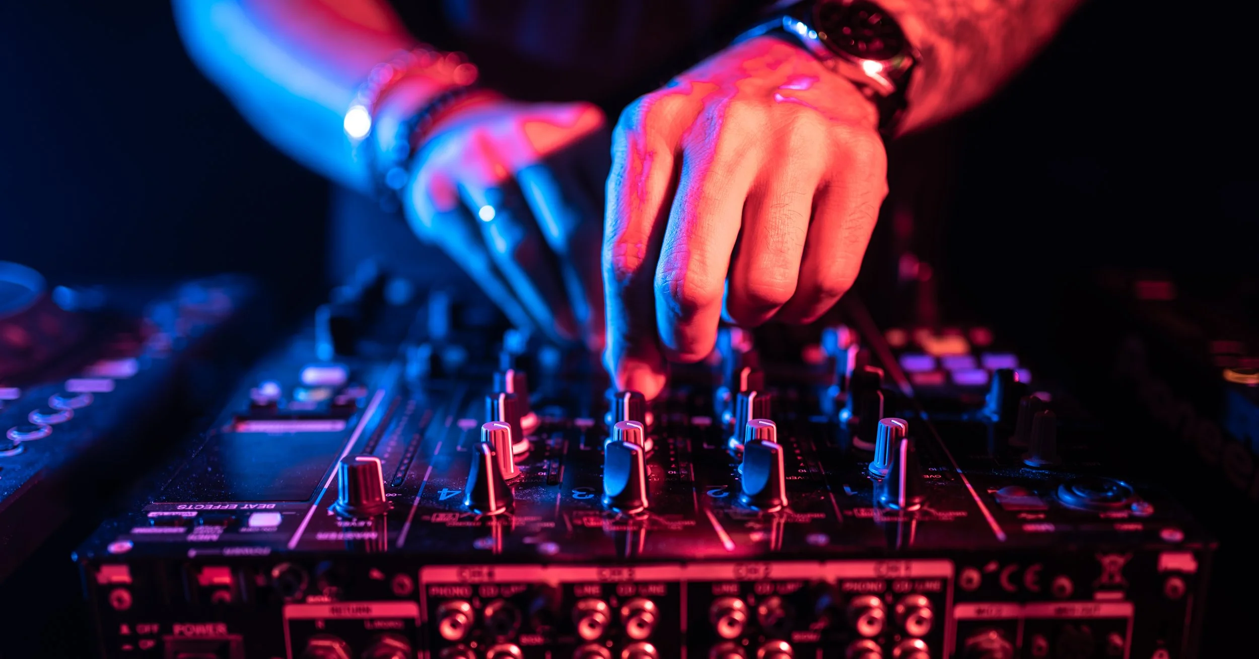 Close-up of a DJ's hand adjusting controls on a sound mixer with colorful neon lights reflecting off the equipment.