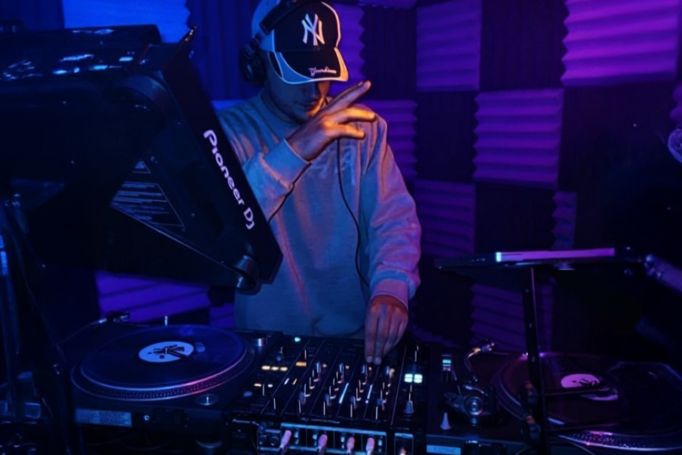 A DJ wearing headphones and a New York Yankees cap plays music on a mixer in a dark, soundproofed room with purple acoustic foam panels.
