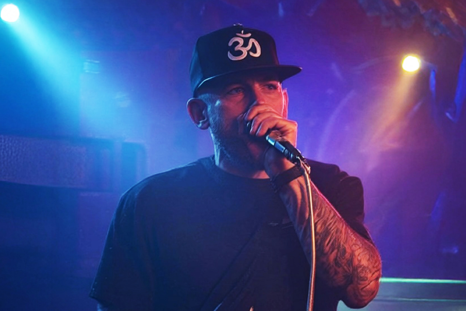 A man singing into a microphone on stage, wearing a black cap with a white Om symbol, and a black shirt, with colorful stage lights in the background.