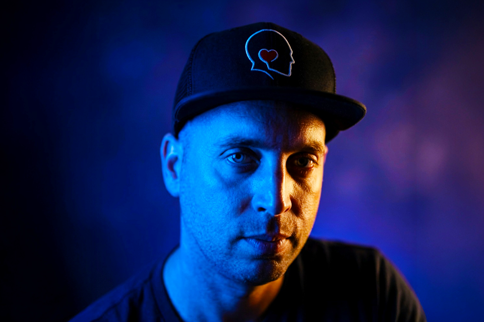 A man wearing a black cap with a heart and brain outline logo, illuminated by blue and purple lighting.