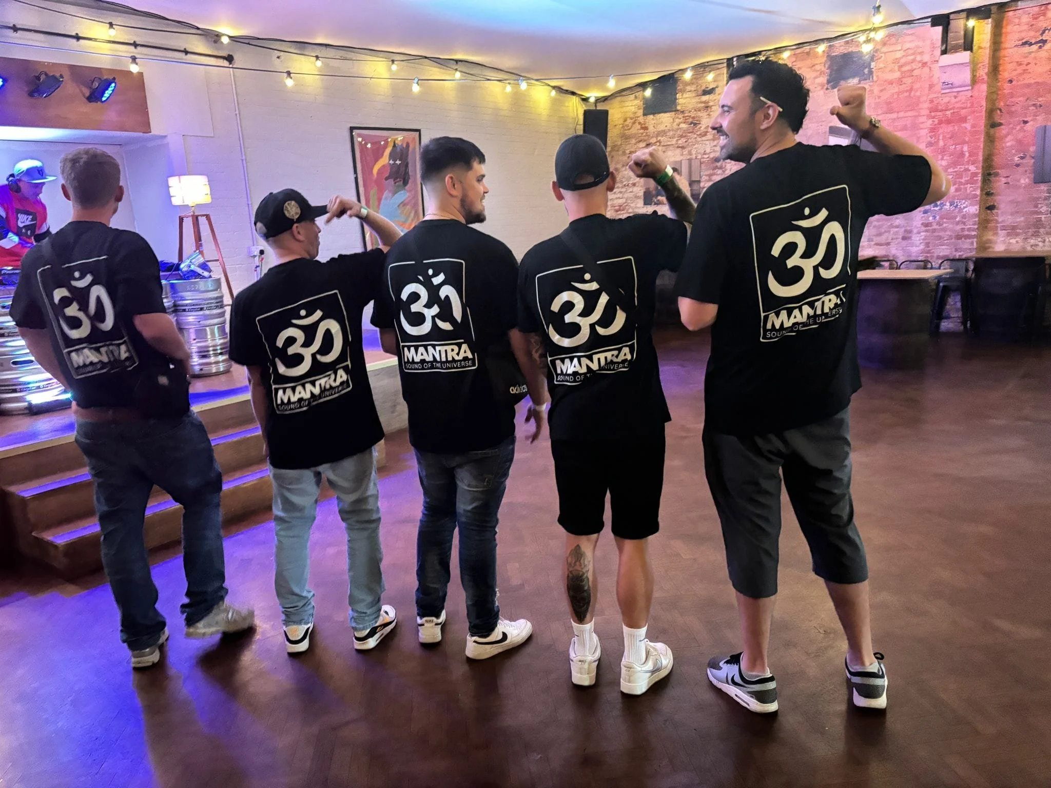 A group of five people standing in a line at a party or nightclub, all wearing matching black t-shirts with a white Om symbol and the word 'MANTRA' printed on them. One person on the far right is dancing or posing with a raised fist. The room has exposed brick walls, warm lighting, and a colorful painting on the wall.