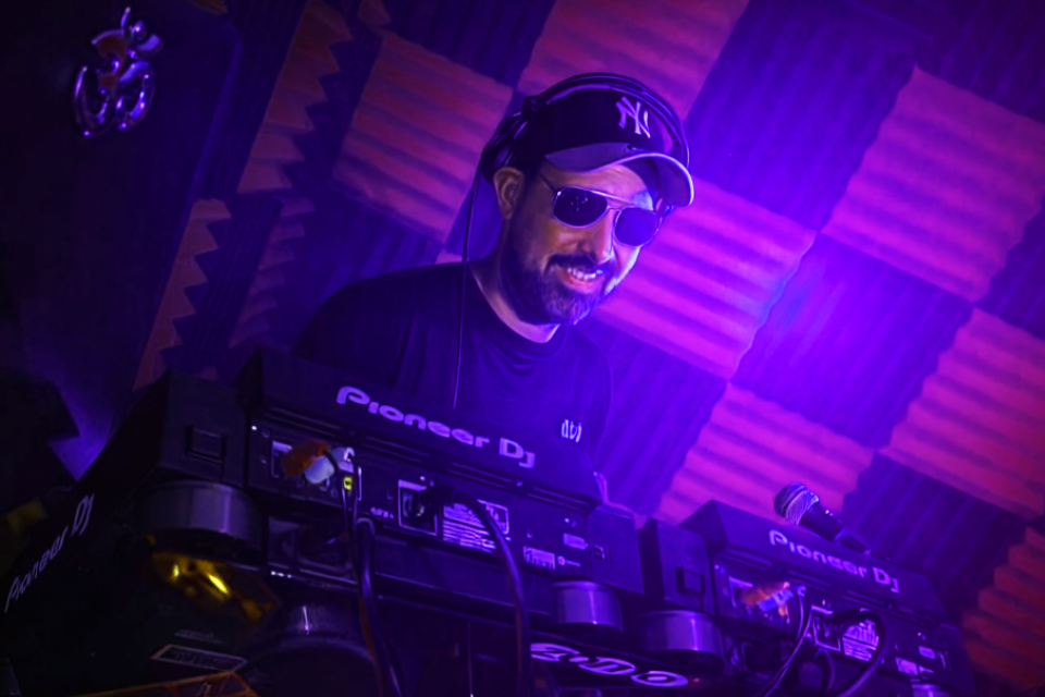 A DJ with a beard, wearing sunglasses, a cap, and a black t-shirt, performing behind a Pioneer DJ controller in a dimly lit room with purple lighting and soundproofing panels.