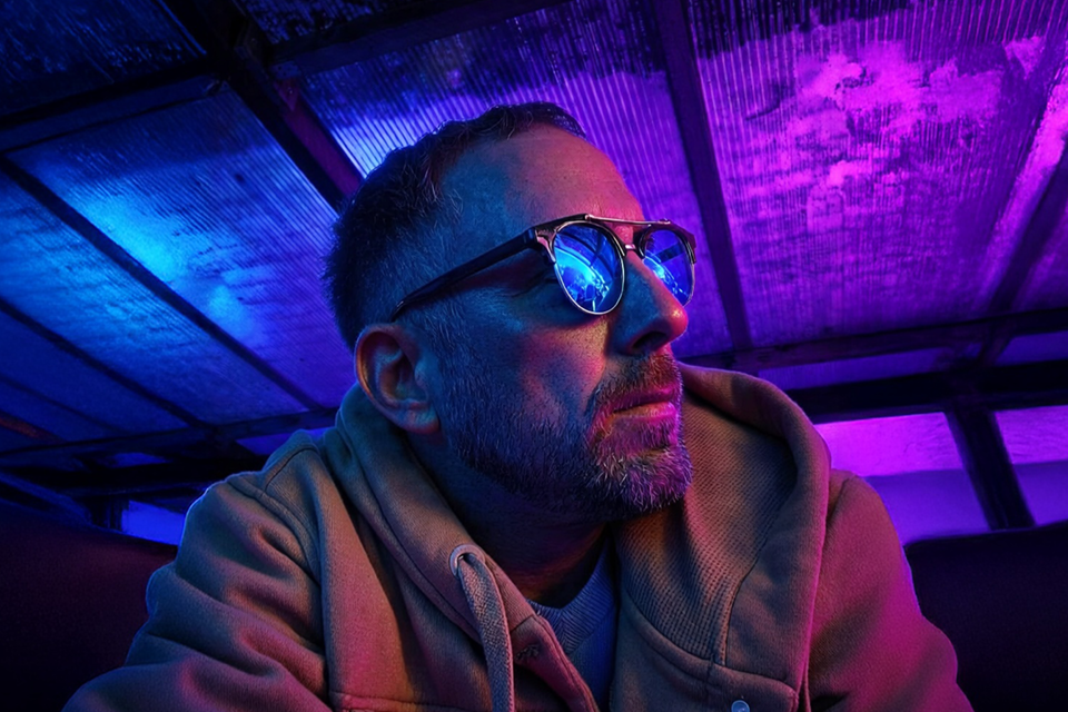 A man wearing glasses with blue neon reflections, in a hoodie, illuminated by colorful purple and blue lighting.