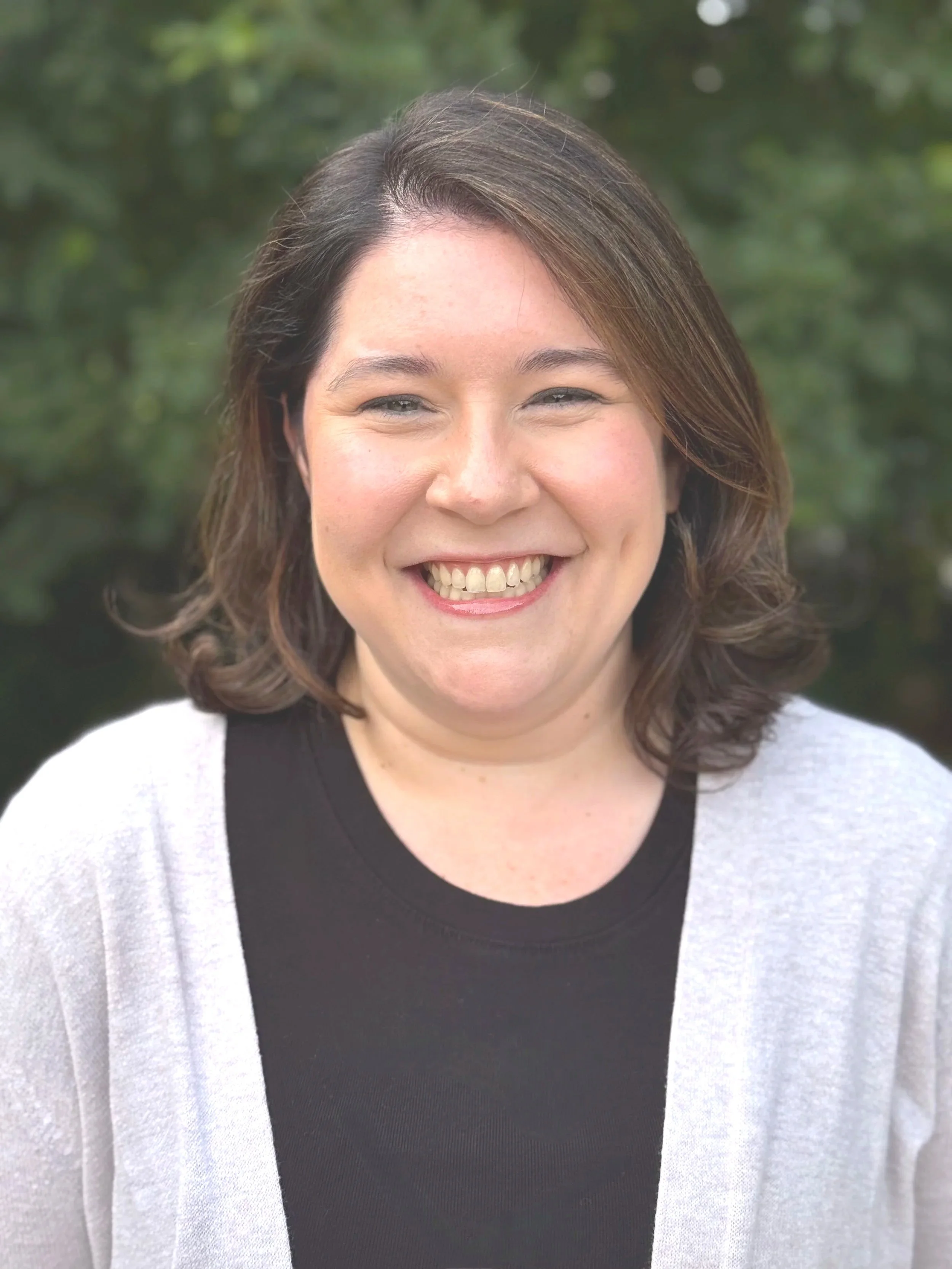 Close-up of a smiling woman with wavy brown hair, wearing a black shirt and a light gray cardigan, outdoors with green blurred foliage background.