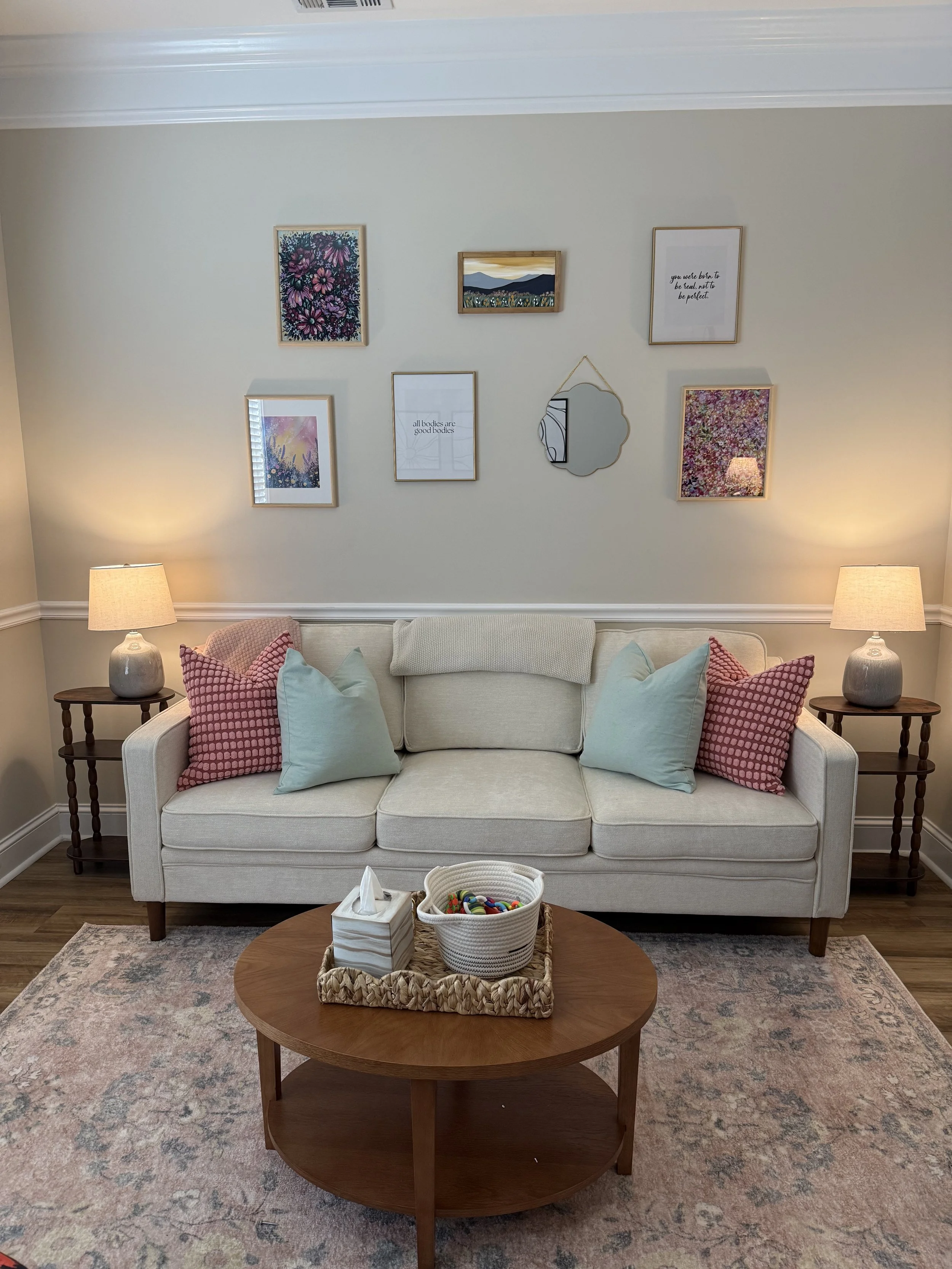Therapy office with a beige sofa, pink and mint pillows, side tables with lamps on each side, a wooden coffee table with tissue box and basket on top, a patterned area rug, and a wall decorated with seven framed pictures and mirror.