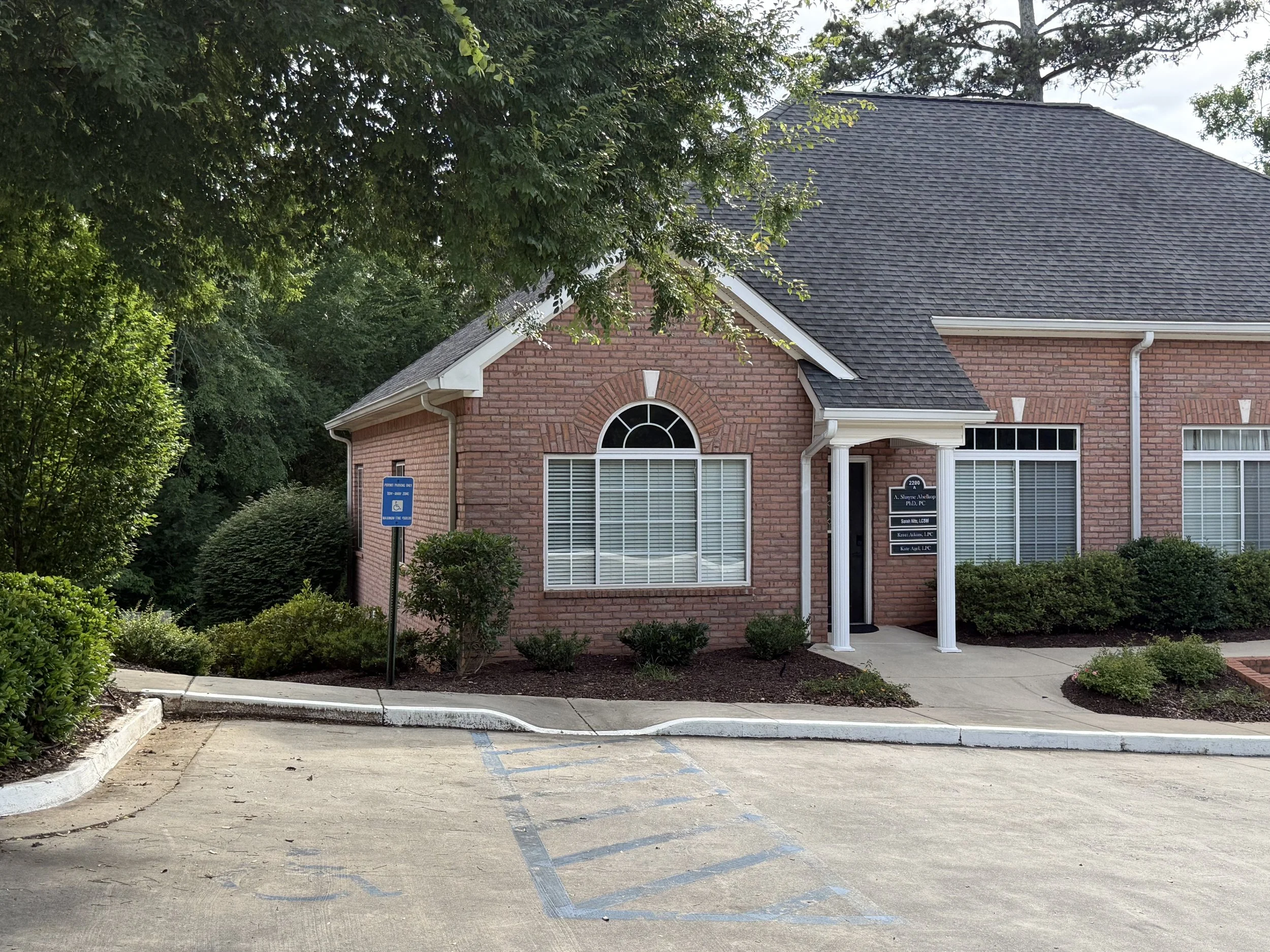 Front view of a brick office building with a curved entrance and large front window, surrounded by greenery including bushes and trees, and a parking lot with reserved parking space marked for handicap parking.