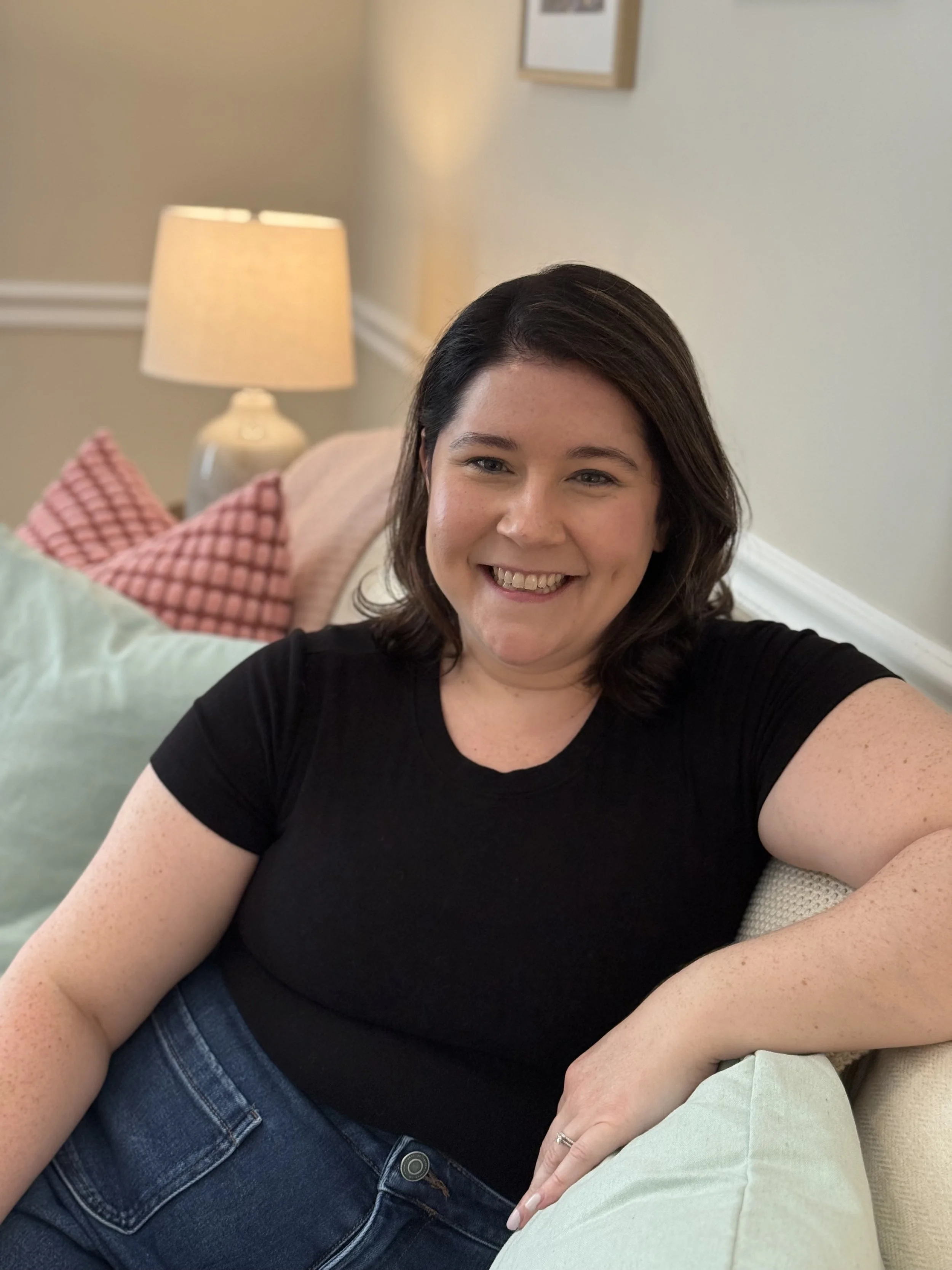 A woman smiling, sitting on a beige couch in a therapy office with a lamp and framed artwork on the wall.