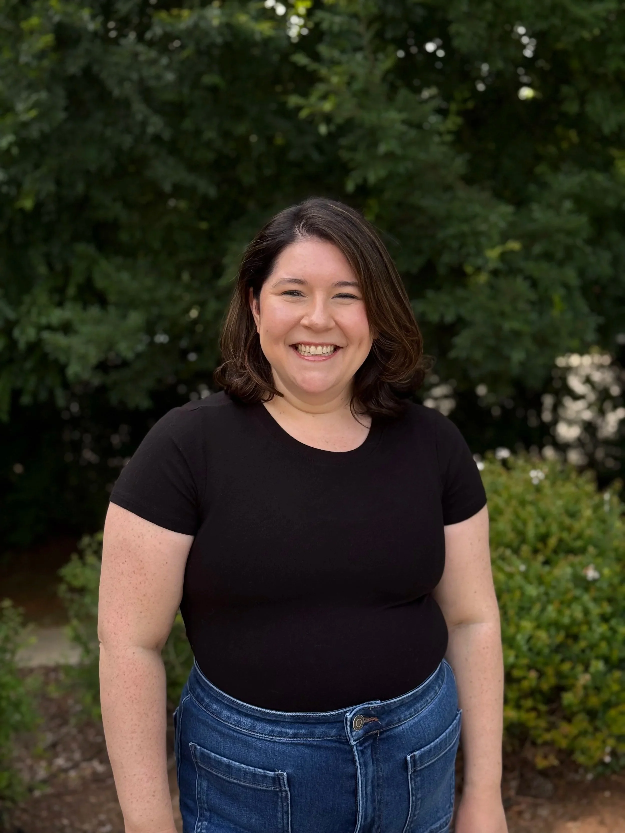 A woman smiling outdoors in front of green leafy trees, wearing a black top and blue jeans.