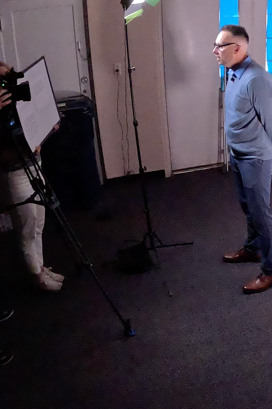 A man in a blue shirt and glasses stands facing a person behind a camera and clipboard. The setting appears to be indoors with professional lighting equipment and plain walls.