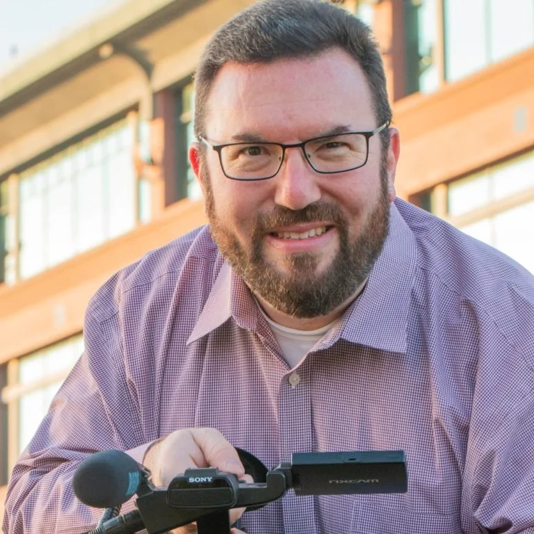 A man with glasses and a beard smiling outdoors, holding a camera, with a modern building in the background.