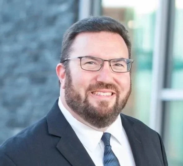 A man with dark hair, glasses, and a beard, wearing a suit and tie, smiling outside a modern building.