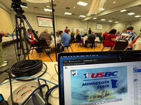 A conference room filled with attendees seated at round tables during a presentation, with a camera setup on a tripod and a laptop displaying the USBC Minnesota State logo in the foreground.