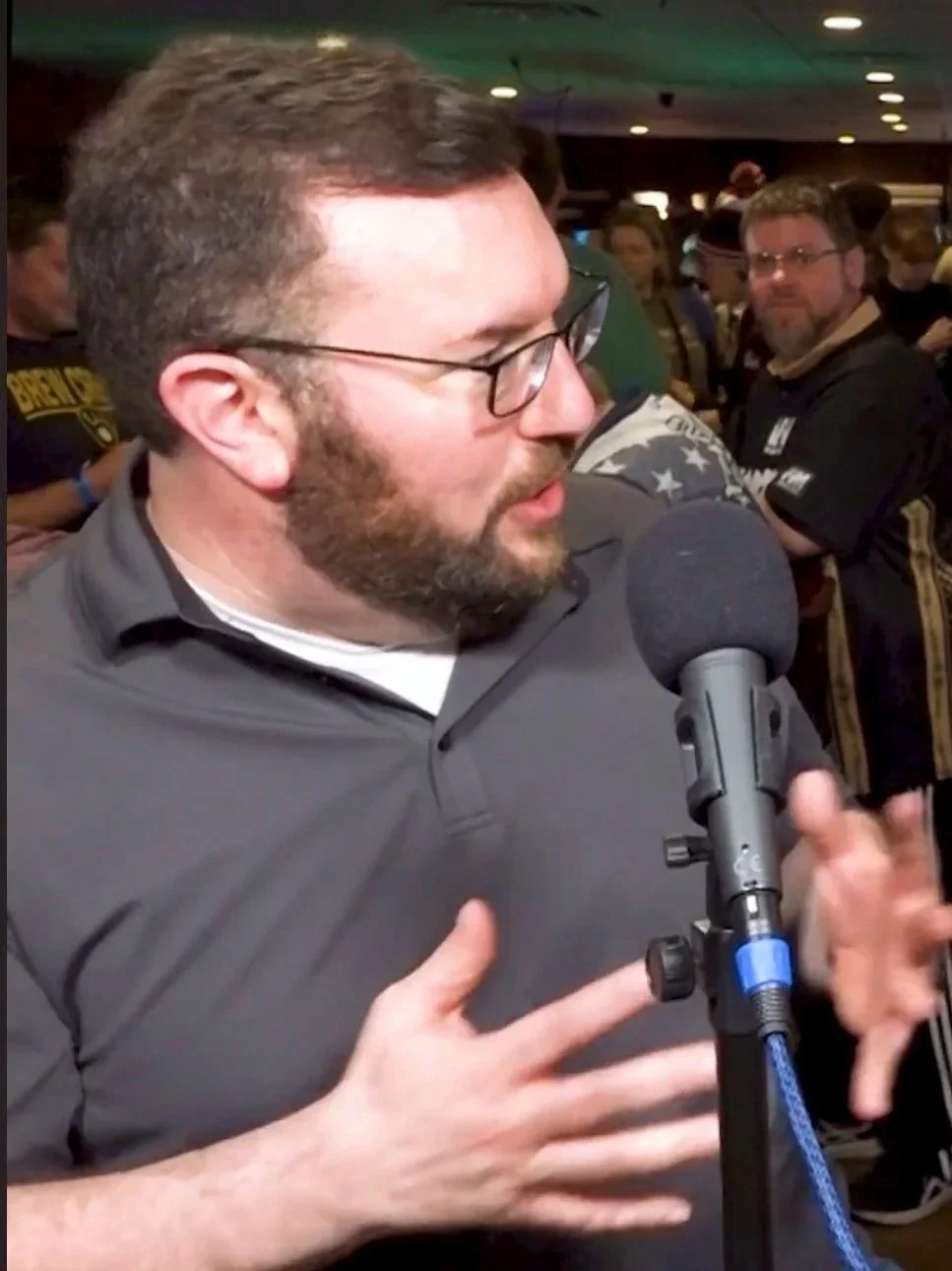 A man with glasses and a beard speaking into a microphone at an indoor event, with other people in the background.
