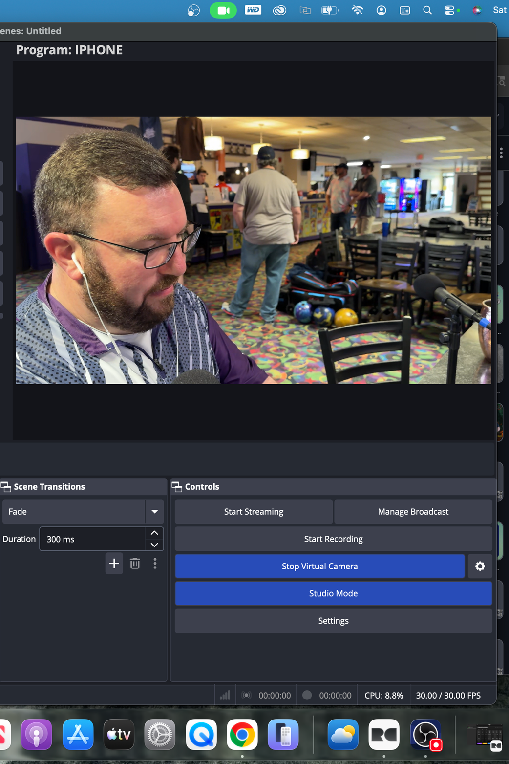 A man with glasses, a beard, and earbuds is seated at a table in a bowling alley or entertainment venue. There are multiple bowling balls and a bag on the floor behind him, with several other people in the background near a counter, colorful carpet, and arcade machines.