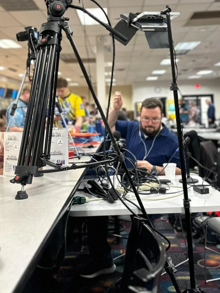 A man sitting at a table in a busy indoor setting, working with electronic equipment and wearing earphones. The photo is taken through a large camera tripod with various wires and gadgets attached.