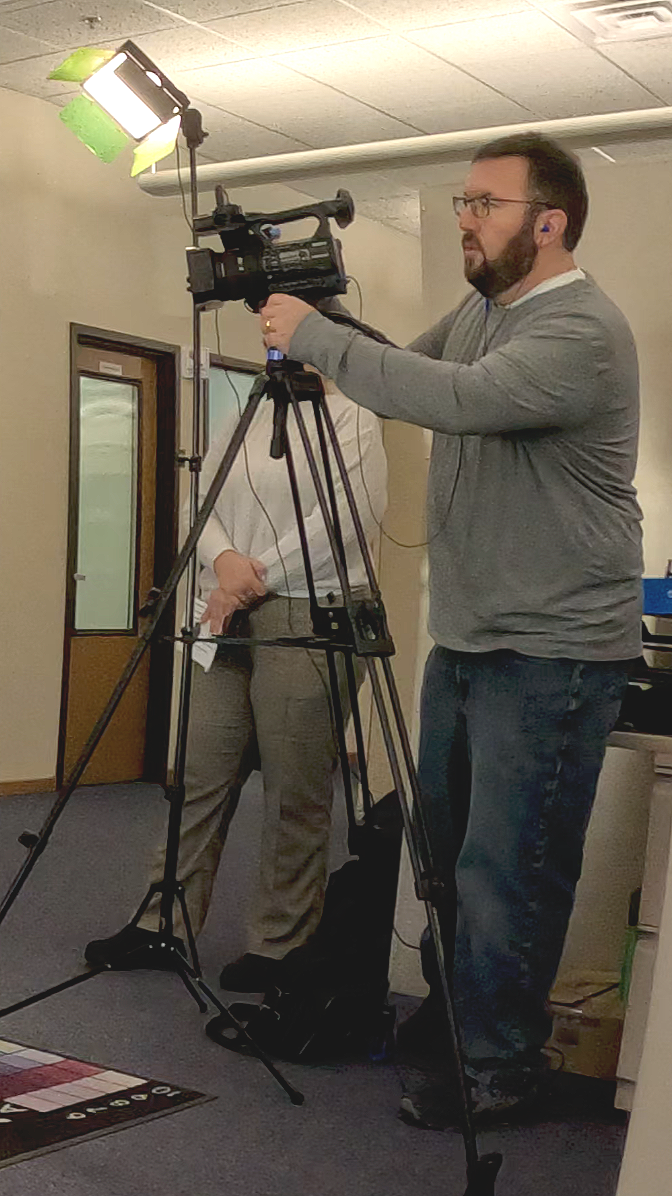 A man with glasses and a beard operating a professional video camera on a tripod, with a bright light attached, indoors.