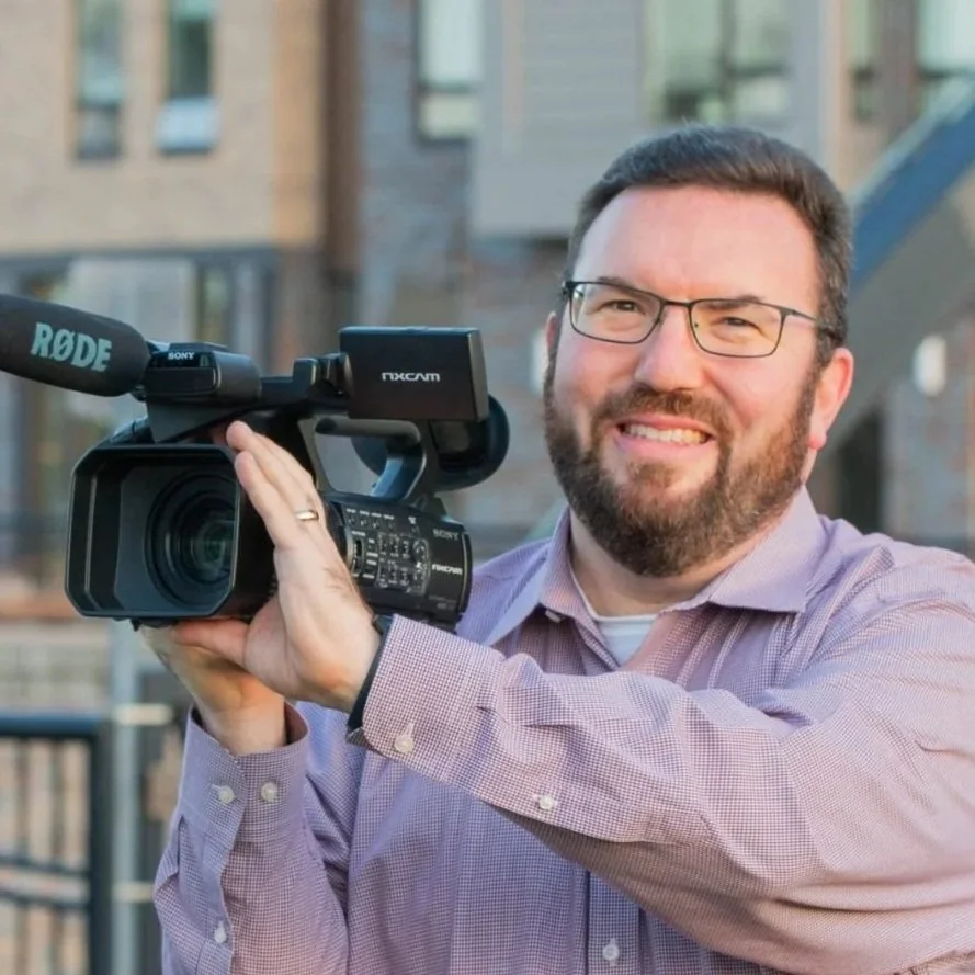 A man with glasses and a beard holds a professional video camera outdoors.
