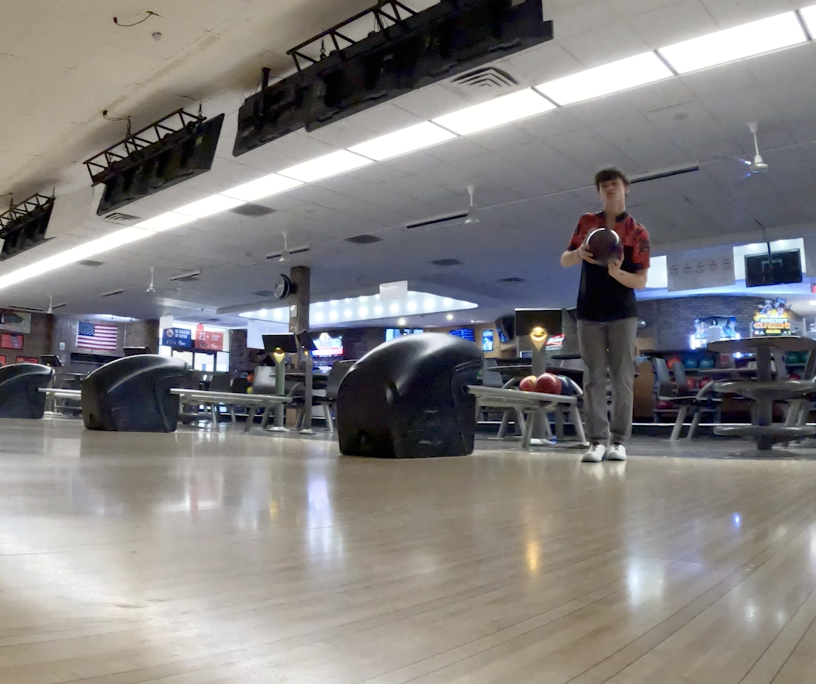 A person standing at a bowling alley holding a bowling ball, with bowling balls and seating visible in the background, and the ceiling with lights and fans overhead.
