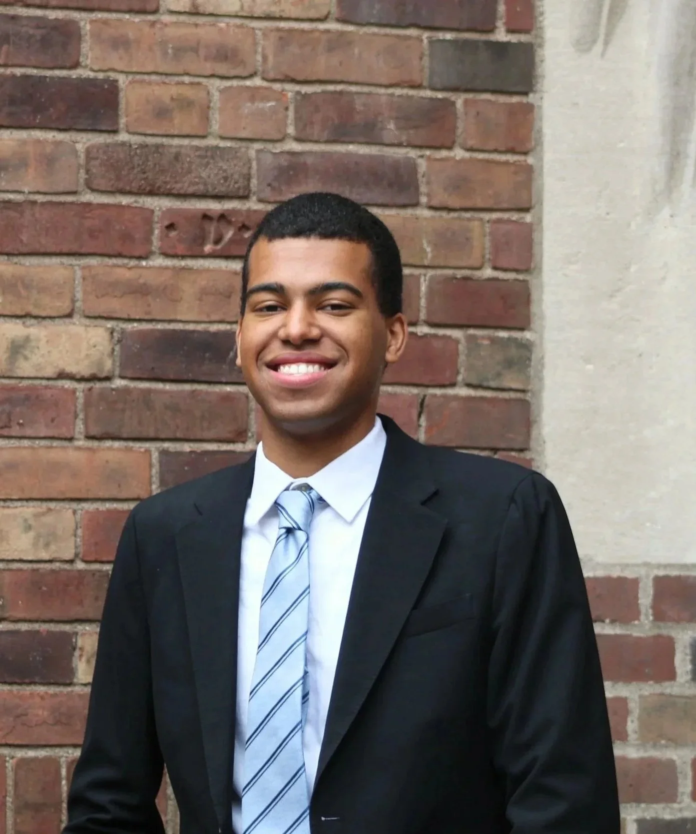 A young man in a black suit and tie standing against a brick wall, smiling.