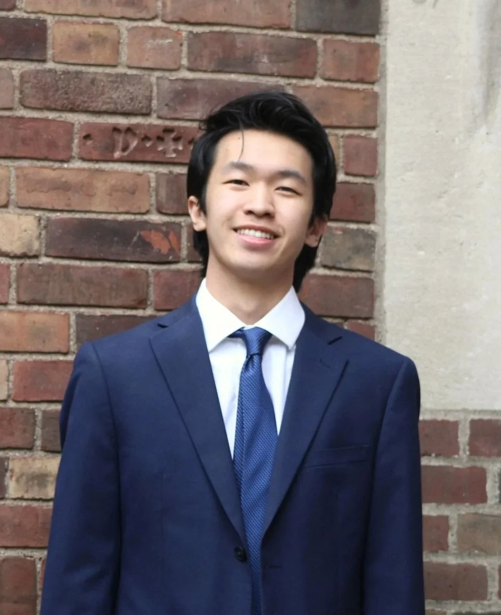 A young man with black hair smiling, wearing a navy blue suit and tie, standing against a brick wall.
