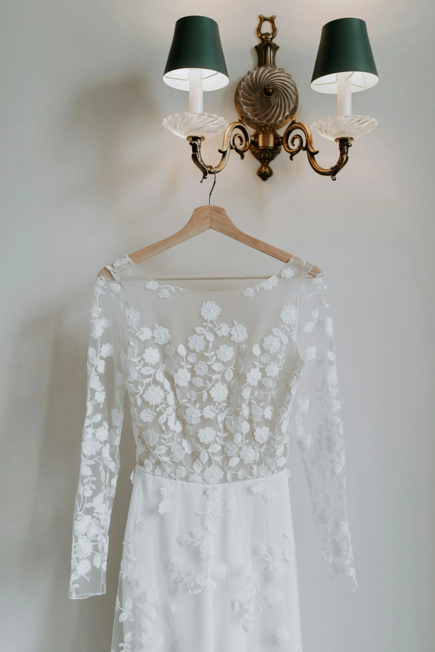 White wedding dress with floral lace top hanging on a wooden hanger under a dual-lamp wall sconce.