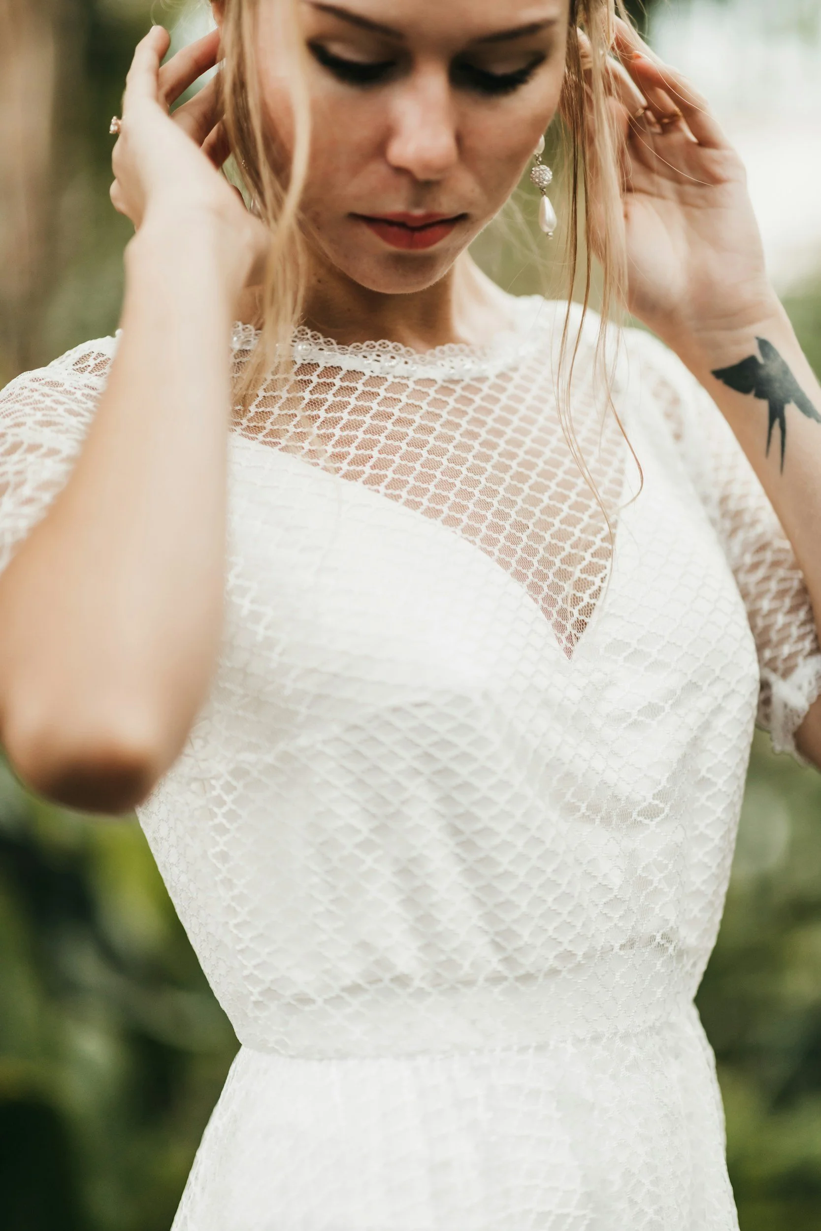 A woman with blonde hair wearing a white lace dress and pearl earrings, adjusting her hair outdoors.