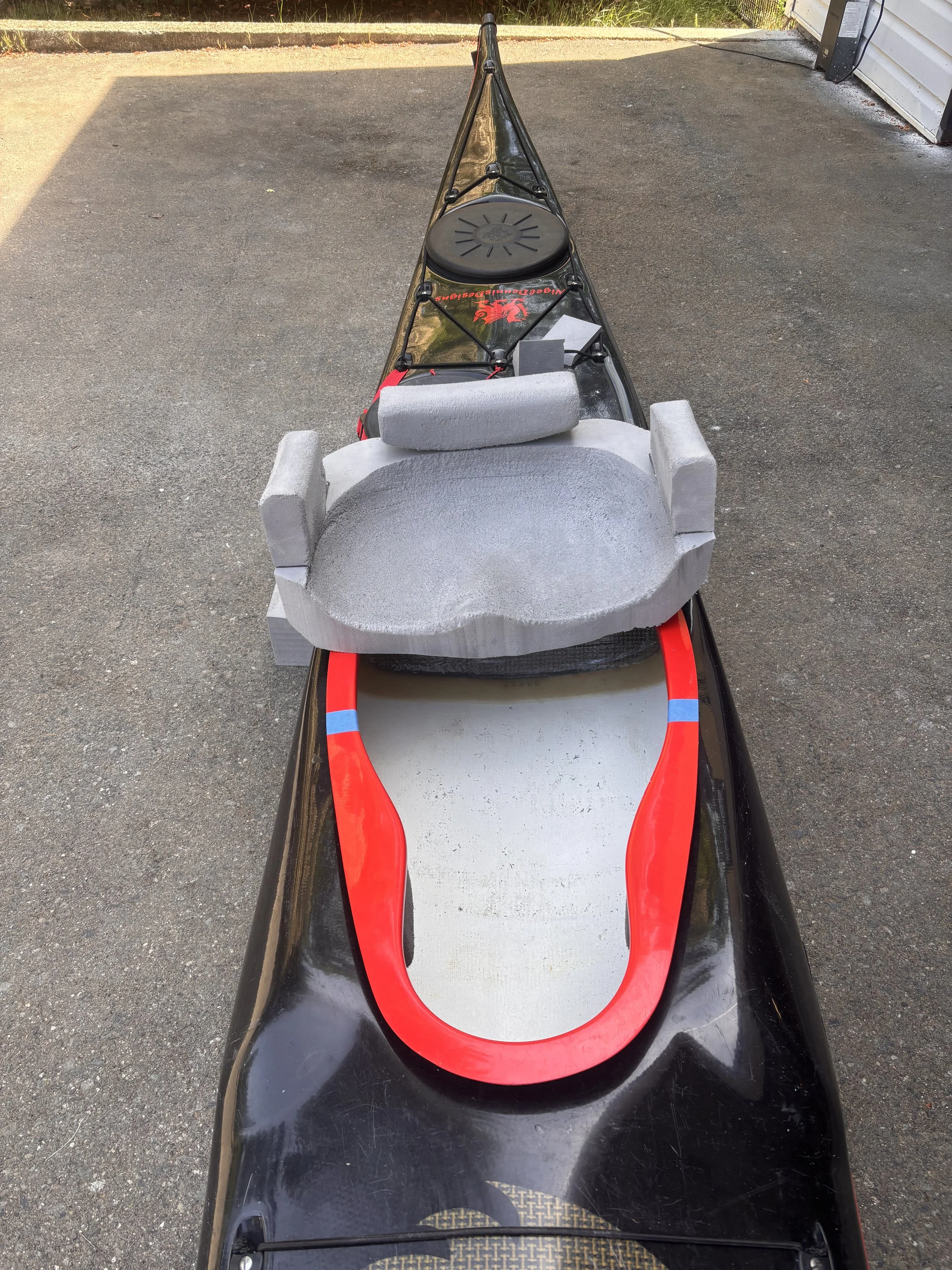 Top view of a black sit-on kayak with a red stripe and a gray foam seat, parked on a paved surface outside a building, with some greenery in the background.