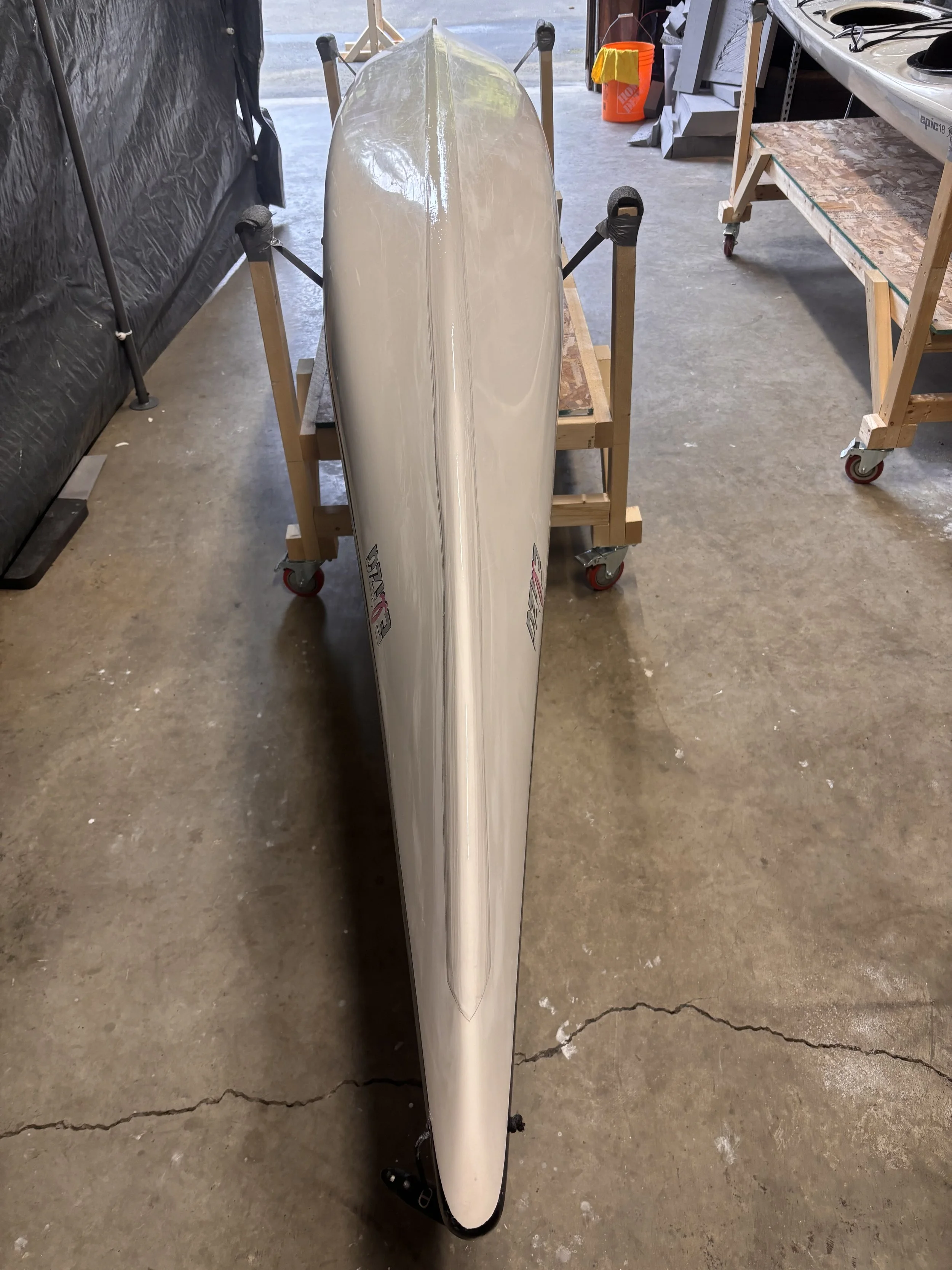 A windsurfing board standing vertically on a wooden stand in a workshop or garage setting.